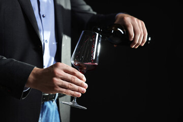 Man pouring red wine from bottle into glass on black background, closeup. Space for text