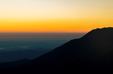 Sunrise over mountain silhouettes with a vibrant gradient sky transitioning from dark orange to pale yellow, creating a serene and tranquil atmosphere