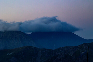 Clouds hovering over dark mountain peaks at dusk, creating a moody and mysterious atmosphere with soft pastel tones in the sky