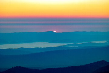  Sunrise emerging over distant mountains, casting a colorful gradient of yellow, orange, and pink across the sky, with layers of blue hills and valleys fading into the horizon