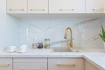 Luxurious kitchen in modern apartment with ecru furniture, marble glossy tiles on the wall and kitchen sink with gold faucet. Real estate.