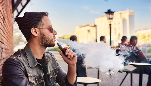 young man vaping relaxed and releases smoke coming out; outside at a cafe.