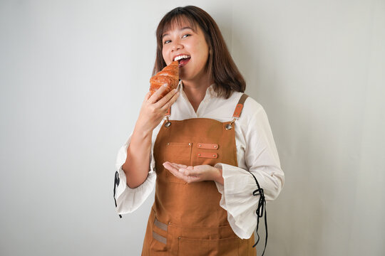 Excited young asian woman wearing long white sleeve shirt and brown apron is eating butter croissant bread with smiling face expression. Isolated over white background.