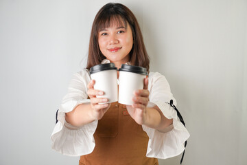 Excited young asian woman wearing long white sleeve shirt and brown apron is holding and serving coffee drinks to customer with smiling face expression. Isolated over white background.