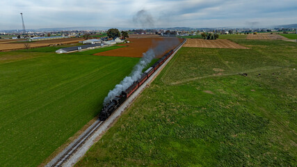 A steam locomotive chugs along the tracks, releasing smoke as it navigates through a scenic rural landscape filled with fields and farms.