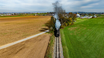 A steam train moves along the tracks beside fields and farms, with smoke billowing into the clear sky on an autumn afternoon.