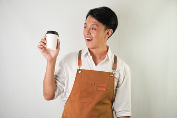 Excited young asian man wearing rolled-up white sleeve shirt and brown apron is holding and serving coffee drinks to customer with smiling face expression. Isolated over white background.