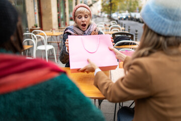 Excited woman receives gift: outdoor cafe surprise, urban friendship, autumn shopping. Joyful expression, pink bag, seasonal fashion, city lifestyle