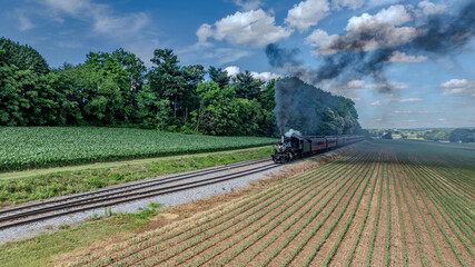 A Black Steam Locomotive Billowing Thick Smoke Pulls Red Passenger Cars Through Lush Green Fields And Forested Areas, Under A Clear Blue Sky With Puffy Clouds, Creating A Picturesque Countryside