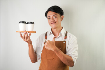 Excited young asian man wearing rolled-up white sleeve shirt and brown apron is holding and serving coffee drinks to customer with smiling face expression. Isolated over white background.