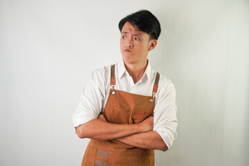 Worried young asian man wearing rolled-up white sleeve shirt and brown apron is thinking about something negative with arms crossed on chest gesture. Isolated over white background.