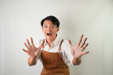 Surprised young asian man wearing rolled-up white sleeve shirt and brown apron with shocked hand gesture. Isolated over white background.