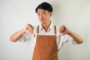 Young asian man wearing rolled-up white sleeve shirt and brown apron with thumbs down hand gesture. Disagreement, bad idea, saying no, rejection. Isolated over white background.