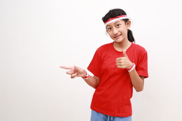 Happy young Asian girl in red white flag ribbon pointing fingers at copy space, showing thumb up gesture isolated on white background. Indonesian independence day on 17 august concept