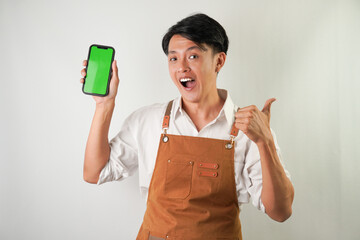 Excited young asian man wearing rolled-up white sleeve shirt and brown apron is showing vertical blank green screen smartphone with thumb up hand gesture. Isolated over white background.