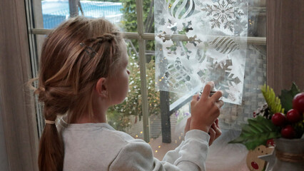 Little girl draw artificial frost through stencil snowflakes on window for Christmas. Decor and...