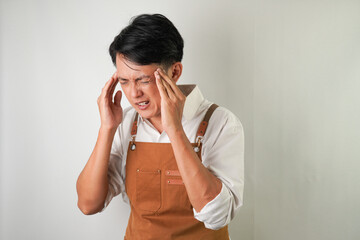 Young asian man wearing rolled-up white sleeve shirt and brown apron is suffering headache and...