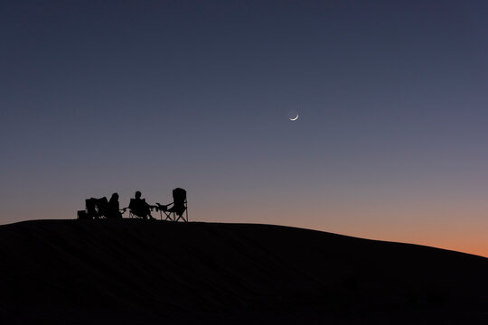 Silhouettes of 2 people sitting on camping chairs on top of a dune enjoying an aperitif at sunset with a moon shining in the orange blue sky Silhouettes of two people on camping chairs atop a dune