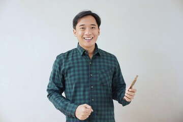 Adult Asian man in green long sleeve flannel shirt looking and smiling at camera while left hand holding smartphone and right hand making fist. Happy, cheerful and satisfaction expression.