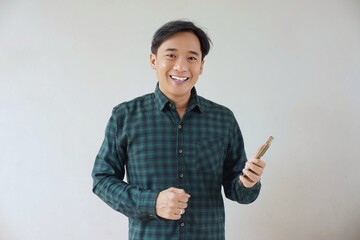 Adult Asian man in green long sleeve flannel shirt looking and smiling at camera while left hand holding smartphone and right hand making fist. Happy, cheerful and satisfaction expression.