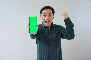 Adult Asian man in green long sleeve flannel shirt holding and showing smartphone display with green screen to camera with right hand while left hand making fist. Happy and cheerful expression.