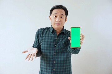 Adult Asian man in green long sleeve flannel shirt holding and showing smartphone display with green screen to camera with left hand while opening right hand palm. Unhappy and unsatisfied expression.