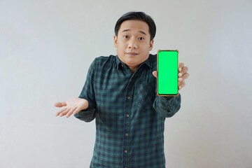 Adult Asian man in green long sleeve flannel shirt holding and showing smartphone display with green screen to camera with left hand while opening right hand palm. Unhappy and unsatisfied expression.