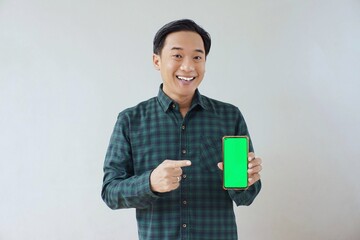 Adult Asian man in green long sleeve flannel shirt showing smartphone display with green screen to camera with left hand while right hand pointing finger to screen. Happy and cheerful expression.