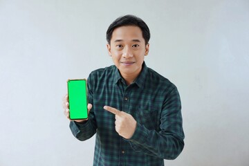 Adult Asian man in green long sleeve flannel shirt showing smartphone display with green screen to camera with right hand while left hand pointing finger to screen. Happy and cheerful expression.