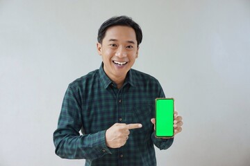 Adult Asian man in green long sleeve flannel shirt showing smartphone display with green screen to camera with left hand while right hand pointing finger to screen. Happy and cheerful expression.