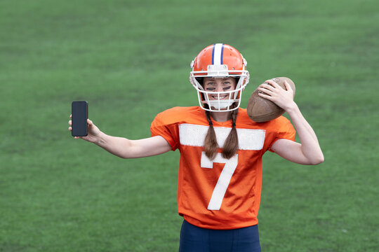 A girl or young woman in a helmet and an American football uniform is holding a ball in one hand and showing a smartphone with the screen facing you in the other. In the background, there is green gra - Powered by Adobe