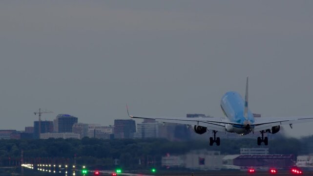White passenger airplane landing on a runway at dusk, approaching the camera with city skyline in the background