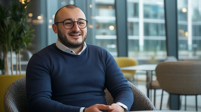 A smiling Middle-Eastern man in a navy sweater sits comfortably in a modern cafe, showcasing a friendly demeanor and stylish eyeglasses.