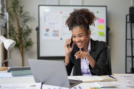 Cheerful young manager is multitasking, talking on the phone while pointing at a laptop screen in a modern office