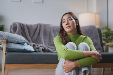 Young woman is sitting on the floor near the sofa, hugging her knees and looking away with a pensive expression
