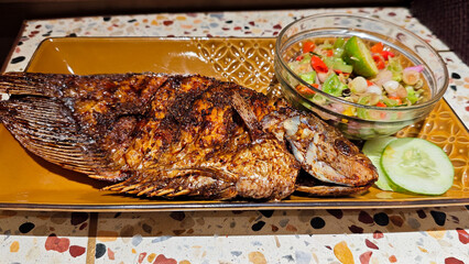 Close-up of Grilled Gourami Fish Served with Balinese Sambal Matah and Fresh Cucumber Garnish on a Traditional Brown Plate in a Restaurant with Terrazzo Table Background