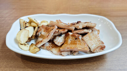 Korean Pork Belly Barbeque Grilled with Roasted Garlic on a White Plate with Light Wooden Table Background