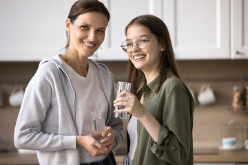 Smiling mature woman and teenage girl stand in kitchen, hold glasses with mineral aqua, care about health together, drink fresh water, quench thirst, lead healthy lifestyle, refreshing on summer day