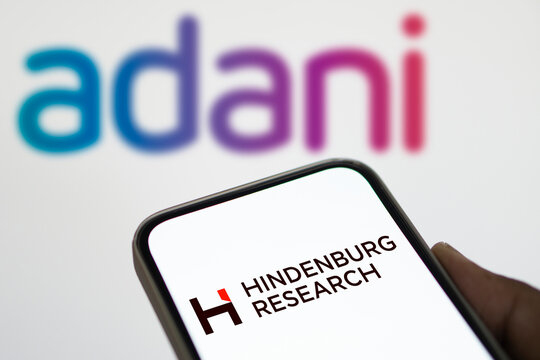 Dhaka, Bangladesh- 18 Aug 2024: The Hindenburg Research logo is displayed prominently on a smartphone screen in the foreground, with the Adani logo blurred in the background.