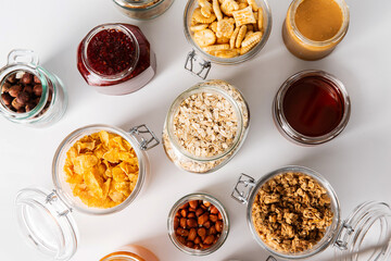 food storage and eating concept - close up of jars with oat, corn flakes, granola, cookies and spreads on white background