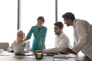 Multi-ethnic young and middle-aged laughing internists using laptop gathered in modern conference...