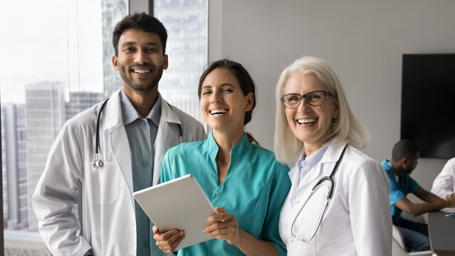 Three multiracial internists team mates looking at camera posing with tablet device standing together in clinic office. Portrait of successful healthcare professional, modern technology usage for work - Powered by Adobe