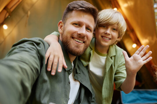 Smiling father and son taking selfie while enjoying camping trip under tent lights during evening, both appearing happy and relaxed - Powered by Adobe