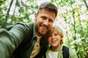 Portrait of father and son smiling for camera while taking selfie in lush green forest showing cheerful moment during outdoor adventure bonding experience