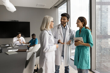Three doctors, Indian, European and Hispanic internists in medical attire standing in modern clinic office, share opinion, skills, insights. Talk between experienced and young healthcare professionals
