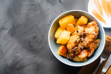 Rustic chicken with potatoes in a blue bowl on a gray background on a gray textured background with a napkin and cutlery. Copyspace.