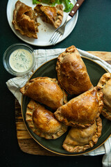 Six delicious freshly baked homemade meat empanadas on green plate served with creamy dip on wooden board on green background.