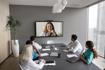 Group of doctors engaged in videoconference with colleague displayed on large screen in conference room, listen and share opinions, discuss medical topics. Remote professional medical virtual meeting