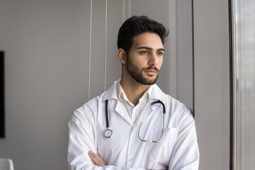 Arabic male doctor standing with arms crossed, wear coat with stethoscope around his neck, look pensively out window, contemplate medical case, diagnosis, or treatment plan. Healthcare worker portrait