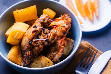 Chicken thighs baked with rustic potatoes with ju-lie sauce on a blue plate on a dark, gray background and a napkin and cutlery.